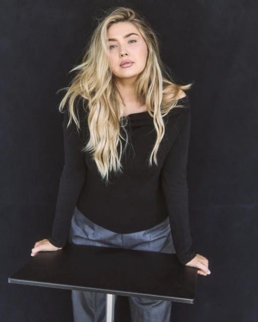 Blonde woman in long-sleeve top poses with hands on a black table against a dark background in a studio portrait shot.