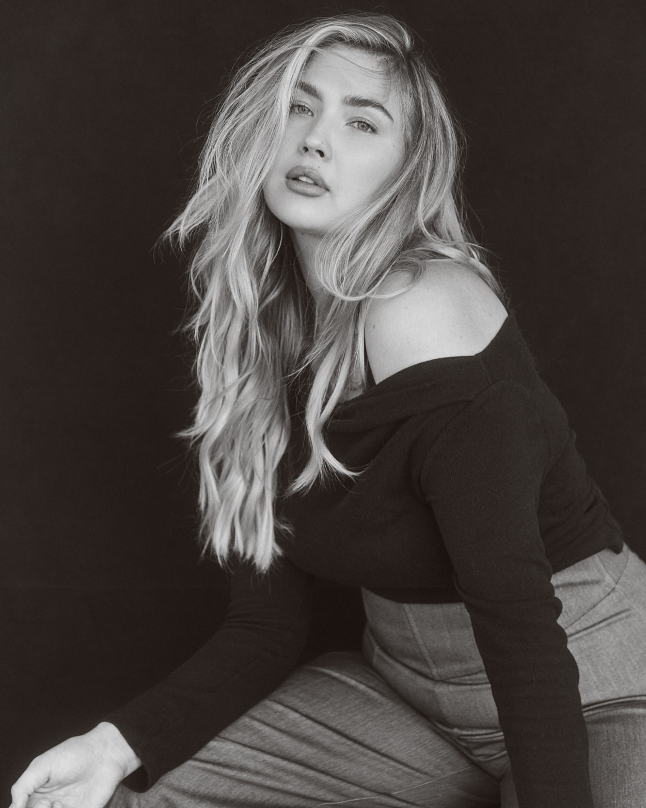 Portrait of a blond adult woman in black top and grey denim, posing indoors against a dark background in a photo studio with soft lighting.