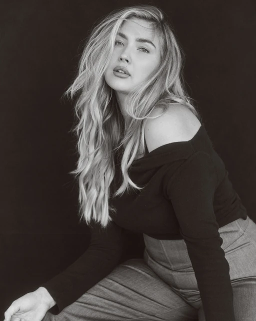 Portrait of a blond adult woman in black top and grey denim, posing indoors against a dark background in a photo studio with soft lighting.