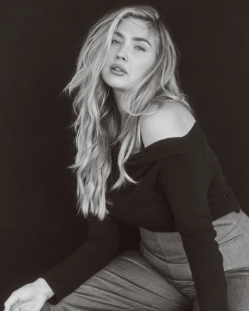 Portrait of a blond adult woman in black top and grey denim, posing indoors against a dark background in a photo studio with soft lighting.