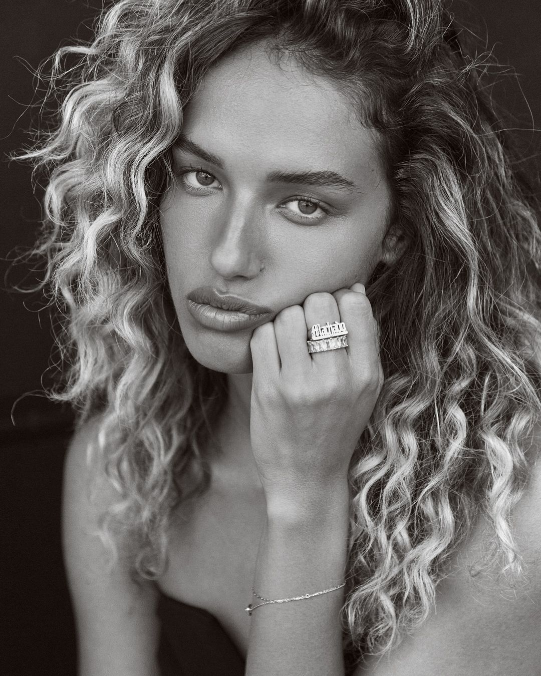 Close-up portrait of a woman with curly hair resting her hand on her chin, displaying two statement rings.