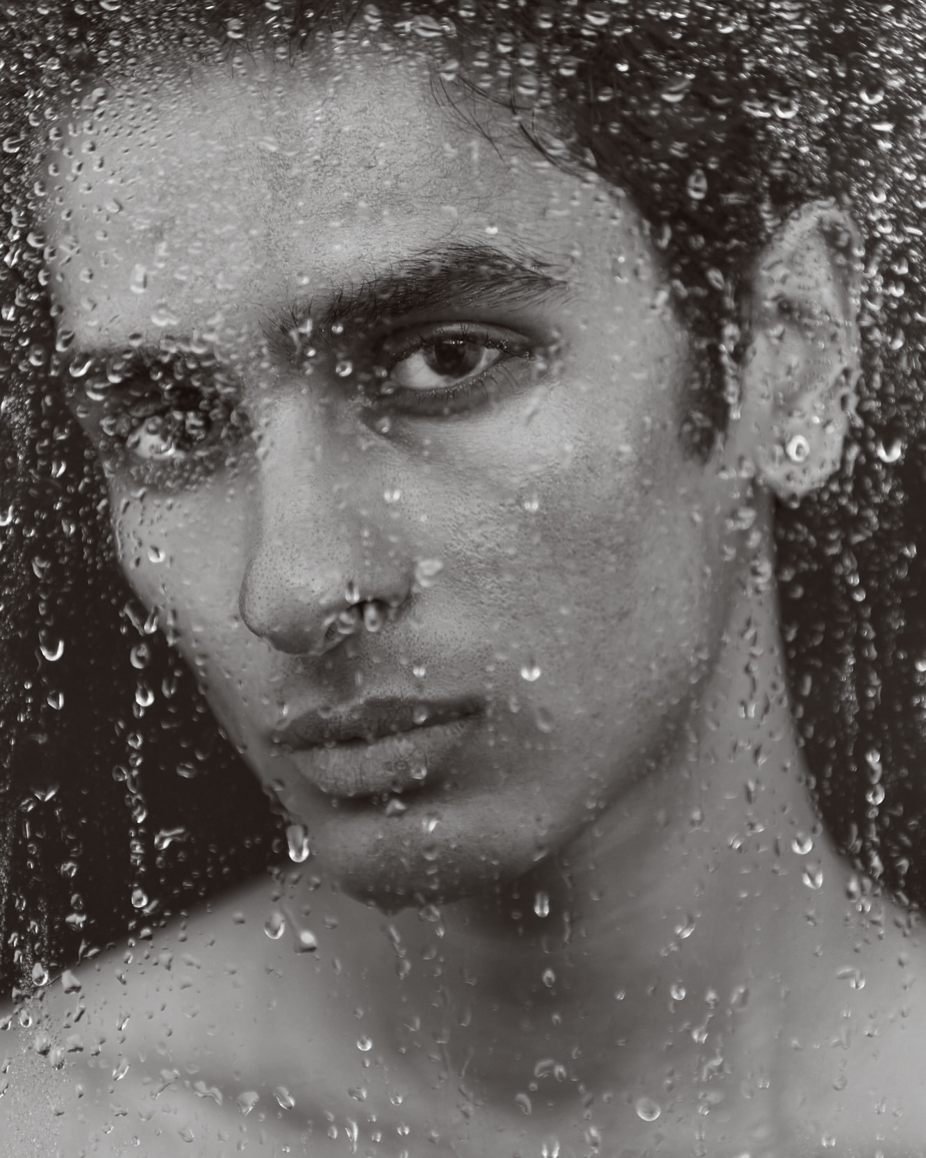 Monochrome portrait of a bare-chested adult male with water droplets on glass, creating a textured, close-up studio shot.
