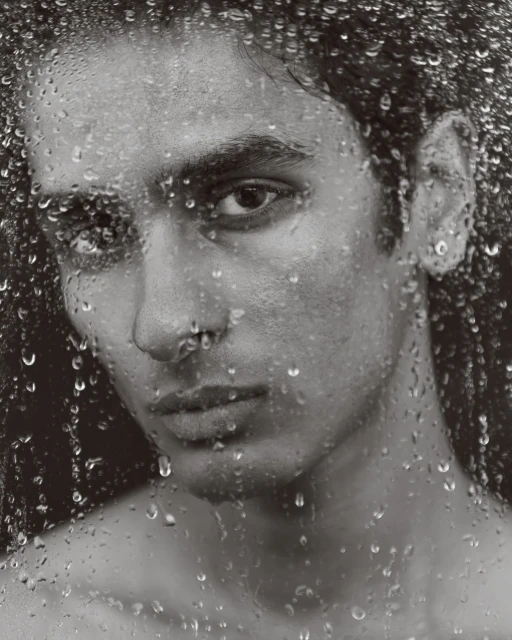 Monochrome portrait of a bare-chested adult male with water droplets on glass, creating a textured, close-up studio shot.