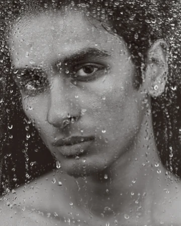 Monochrome portrait of a bare-chested adult male with water droplets on glass, creating a textured, close-up studio shot.
