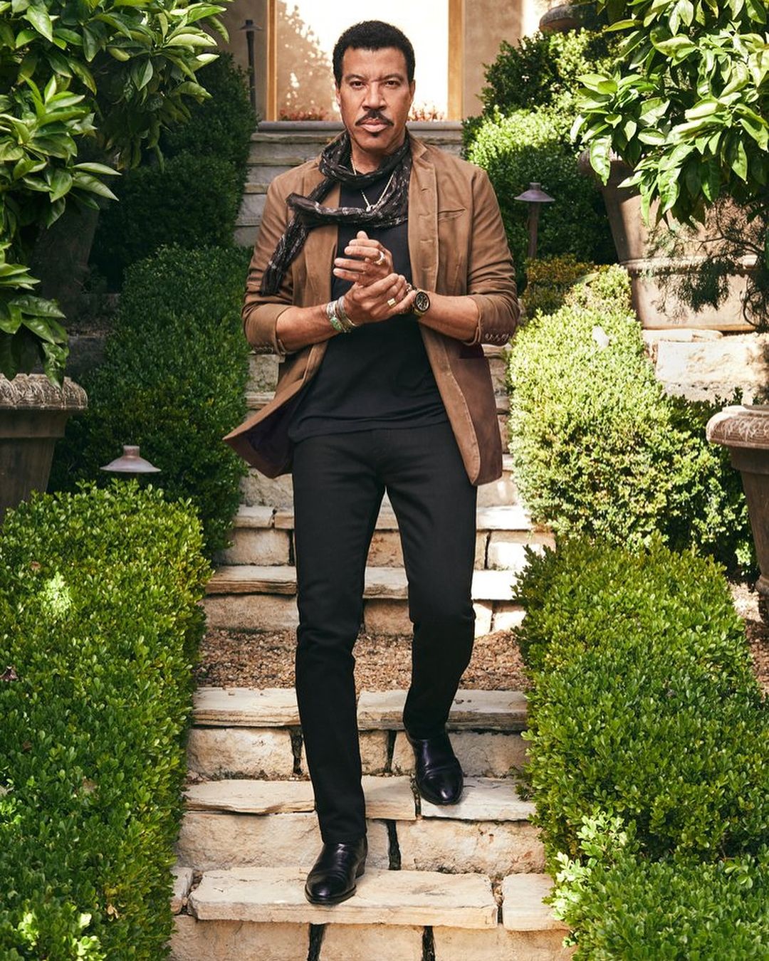 Stylish adult man in a brown jacket, black shirt and pants, and black scarf, walking down stone steps lined by green bushes.