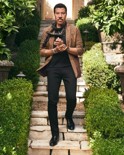 Stylish adult man in a brown jacket, black shirt and pants, and black scarf, walking down stone steps lined by green bushes.