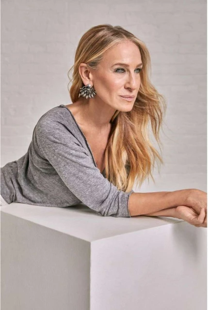 Elegant woman with long blonde hair and statement earrings, wearing a gray long-sleeve shirt, posing against a white background.