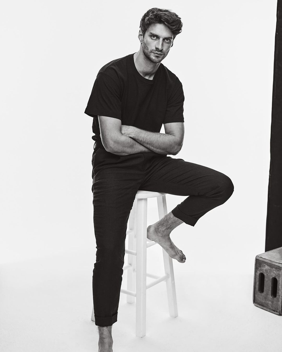 Black and white portrait of an adult male sitting on a stool, looking at the camera with arms crossed in a bright studio environment.