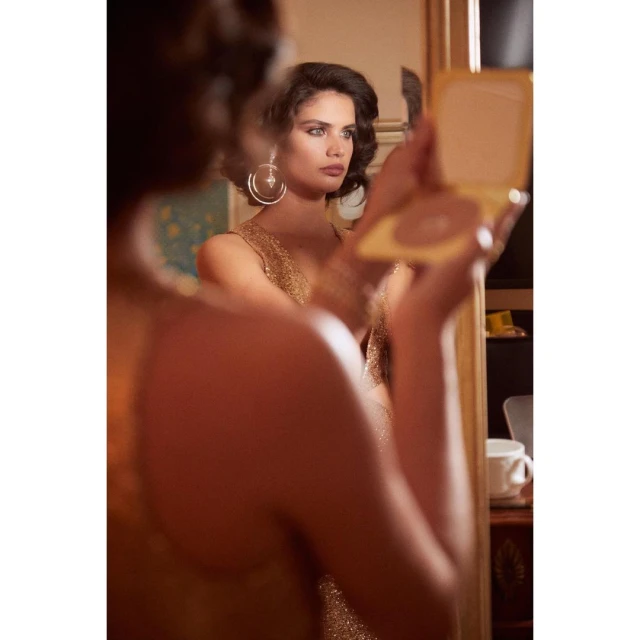 Woman in a gold dress looks in a mirror while holding a compact, indoors with warm lighting. Beauty shot with soft focus.