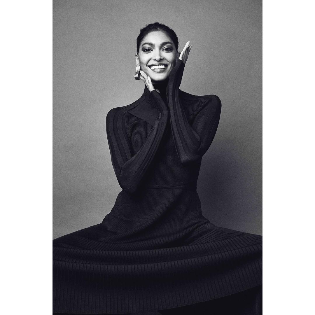 Elegant woman in a black ribbed dress smiles with hands framing her face, creating a striking monochrome fashion portrait.