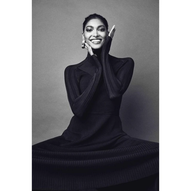 Elegant woman in a black ribbed dress smiles with hands framing her face, creating a striking monochrome fashion portrait.