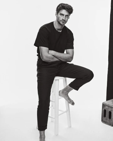 Black and white portrait of a man sitting barefoot on a stool with crossed arms against a white background.