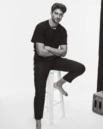 Black and white portrait of a man sitting barefoot on a stool with crossed arms against a white background.