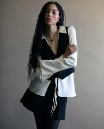 Portrait of a woman with long braided hair wearing a white shirt and black vest against a plain neutral wall