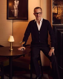 A man in a black cardigan and glasses sitting at a table in a bar with a cocktail glass and small lamp.