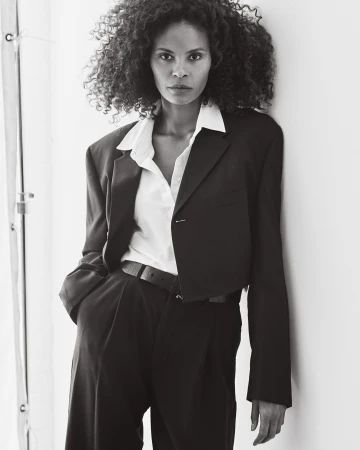 Black and white portrait of a woman with curly hair wearing a white shirt and dark suit jacket leaning against a wall