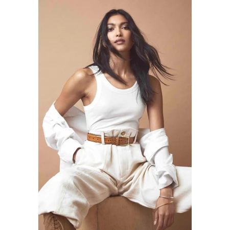 Woman in white tank top and white trousers with brown belt seated against a warm tan background by Andrew Day