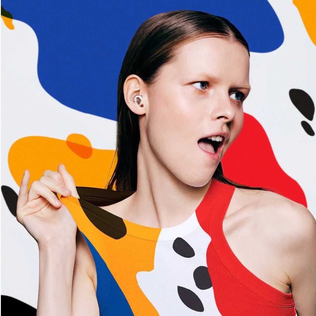 Stylish studio shot of a young adult woman wearing wireless ear buds, bold color block top, with a painterly abstract backdrop.