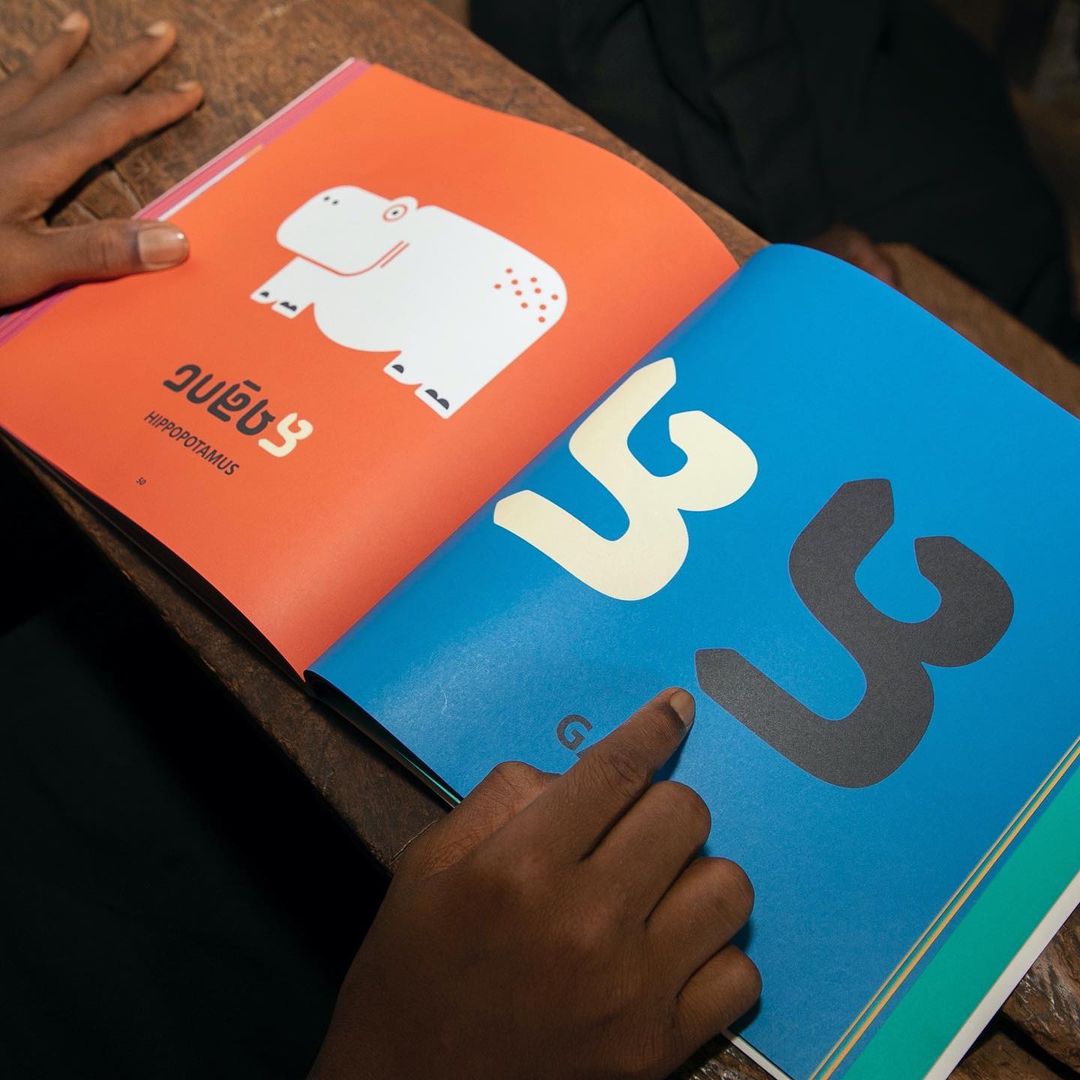 Person pointing at a brightly colored children's book with illustrations of a hippo and the number 3 on a wooden desk indoors.