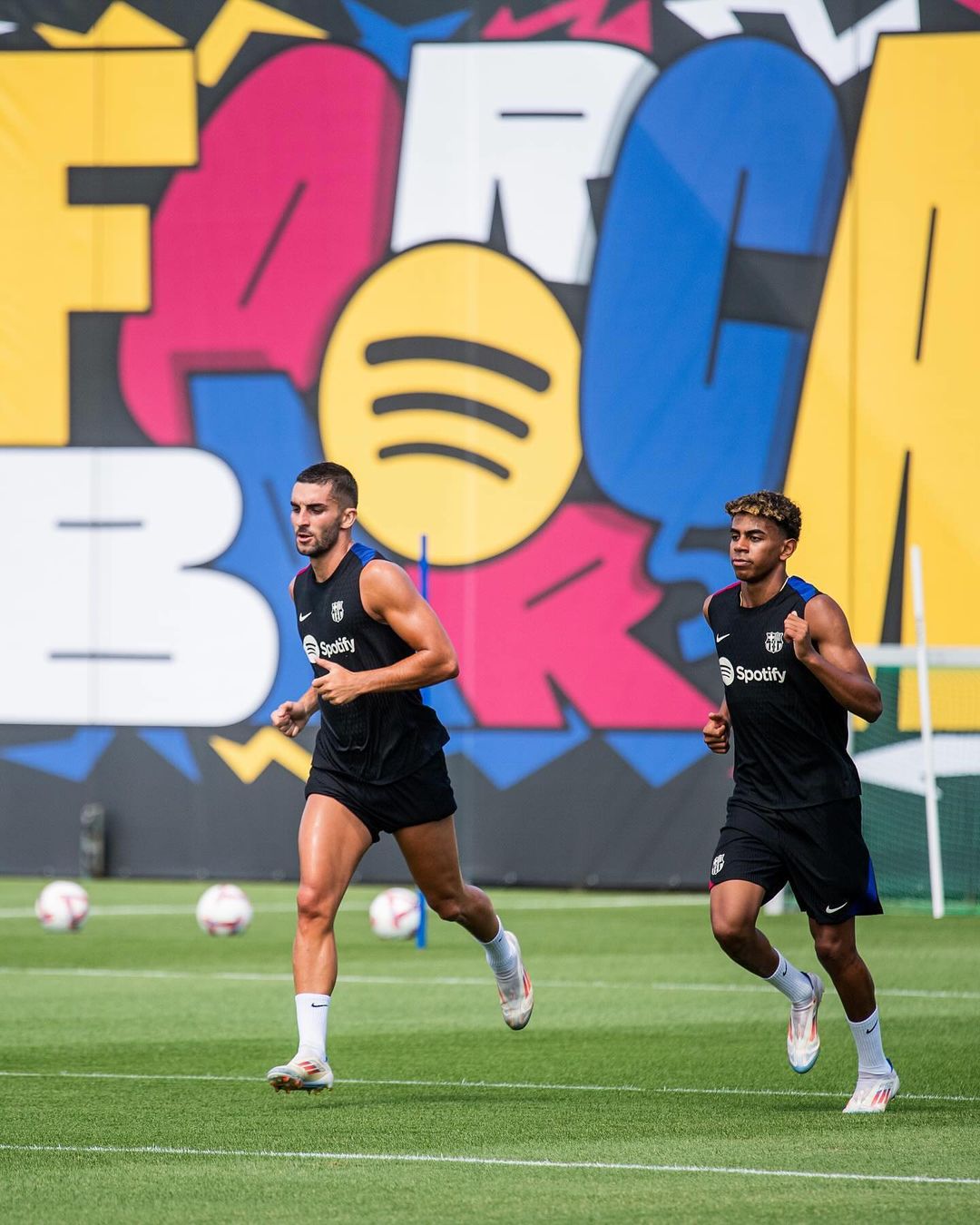 Two athletic men in black tank tops and shorts are running on a green field, with colorful graffiti art on a wall in the backdrop.