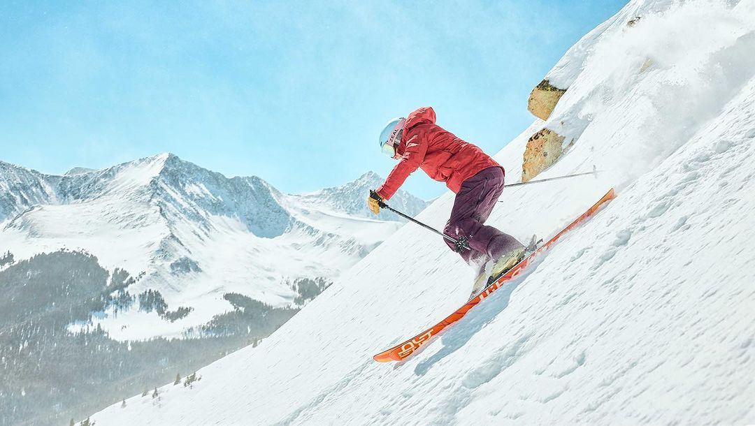 Female skier in red jacket and helmet skiing down snowy mountain slope on sunny day with mountain range in the background.