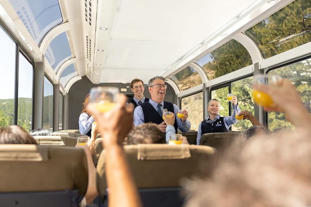 People celebrating with juice on a train with large windows, offering scenic views. Smiling staff are present, adding to the cheerful atmosphere.