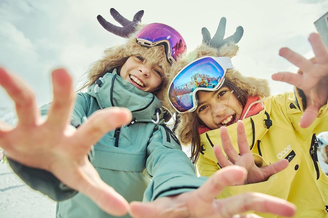 Two smiling children wearing snow goggles and furry hats with antlers in a low-angle shot with outstretched hands on a snowy mountain.