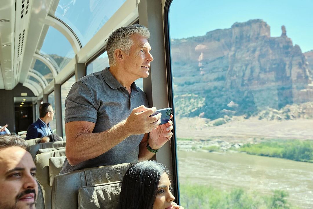 Mature man takes a photo from a train window overlooking a river and canyons, while other passengers sit nearby on a sunny day.