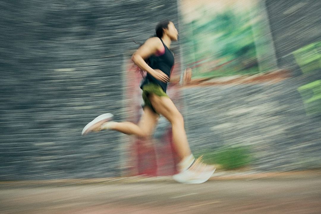 An adult woman is captured in motion, running outdoors with a blurred background suggesting speed and movement, wearing athletic attire and shoes.