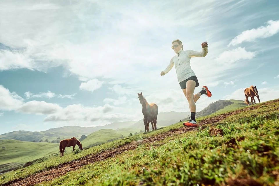 Female runner sprints downhill past grazing horses in a sunny, grassy, hilly landscape with a cloudy sky. Active lifestyle scene.