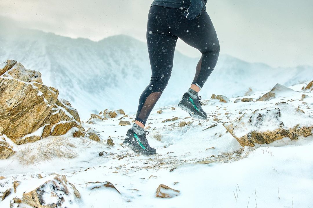 Athlete runs on snow-covered rocky terrain in black leggings and athletic shoes, mountains and overcast sky in the background.