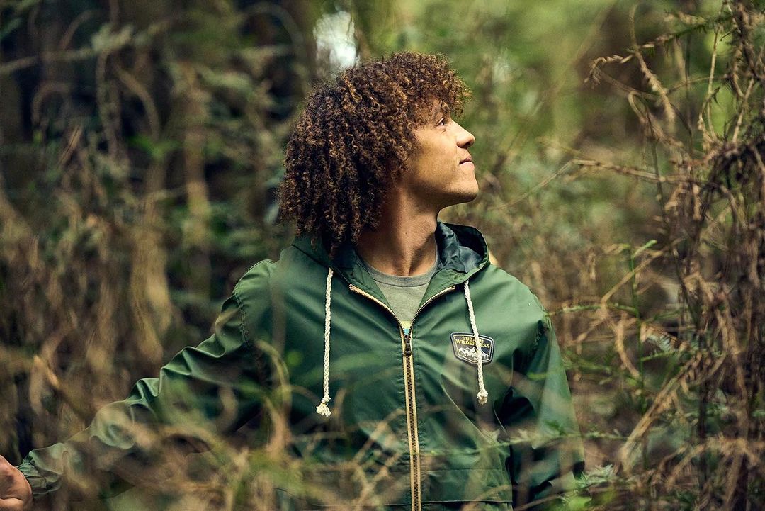 Young adult male with curly hair in a green jacket looks up in an outdoor forest setting with blurred background, medium framing.