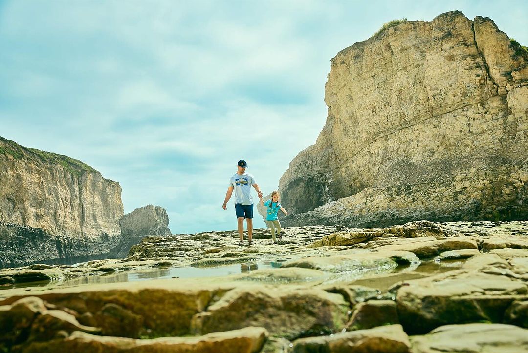 Adult male and child walking on rocky terrain with large rock formations, sunny day, clear sky, puddles on the ground. 