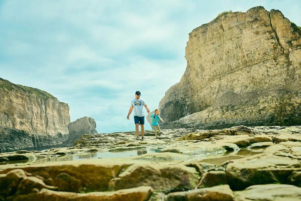 Adult male and child walking on rocky terrain with large rock formations, sunny day, clear sky, puddles on the ground. 