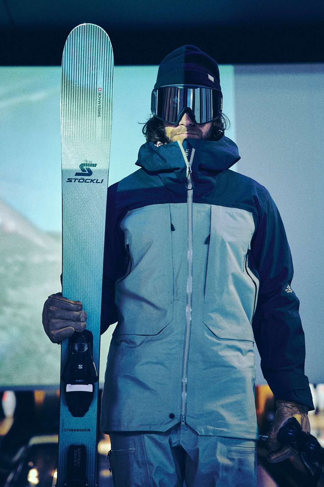 An adult man in ski goggles and a black beanie poses with a ski in a light-filled indoor setting, wearing a two-tone gray jacket.