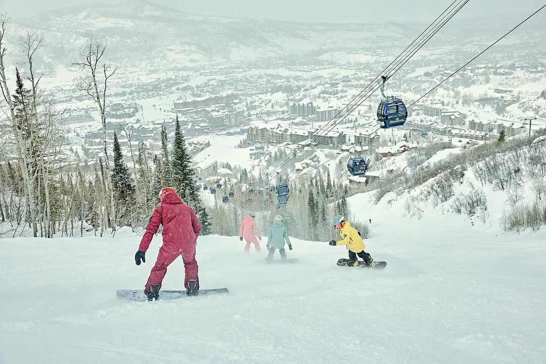 Group of snowboarders ride down a snow-covered ski slope with ski lift and mountains in the background on a cloudy, overcast day.