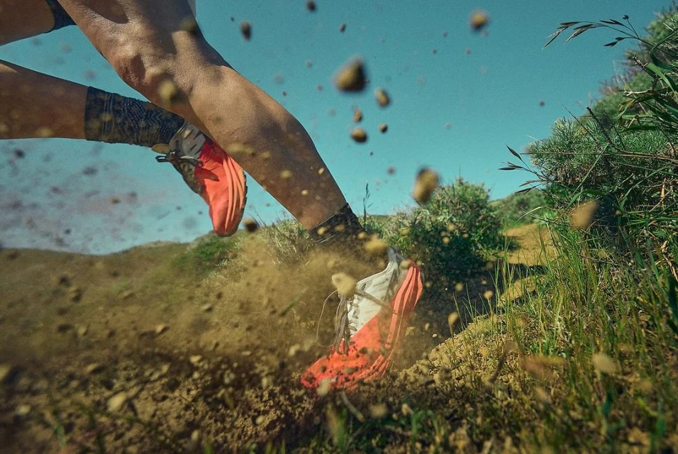 Close-up of a person running on a dirt trail with dirt particles flying around their red and white athletic shoes, set against a blue sky.