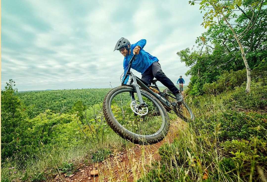 Man mountain biking downhill on a dirt trail with lush trees, wearing a helmet and blue shirt on a cloudy day, action shot.
