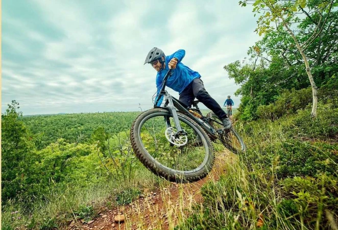 Man mountain biking downhill on a dirt trail with lush trees, wearing a helmet and blue shirt on a cloudy day, action shot.