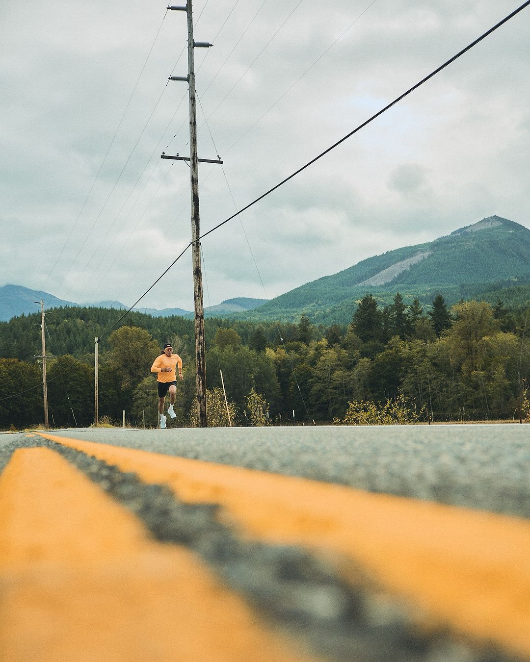 An adult male running on an asphalt road with yellow lines, set against a backdrop of mountains and a cloudy sky.