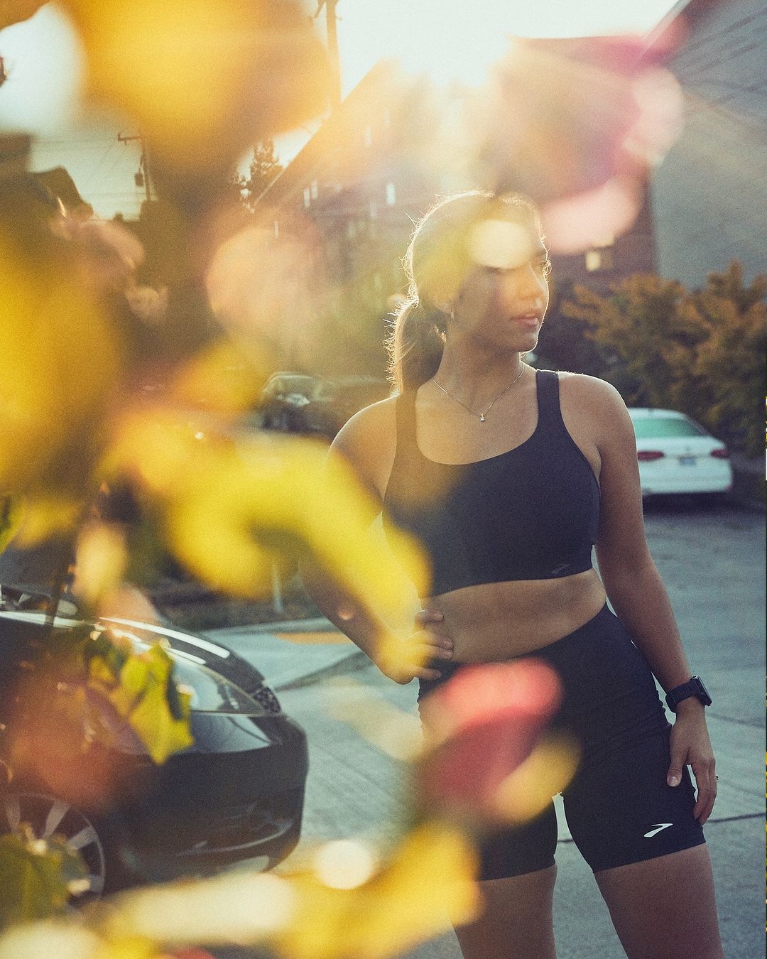 Woman in athletic wear stands outside near cars in filtered sunlight; fitness, healthy lifestyle, sunlight, outdoors, urban setting.