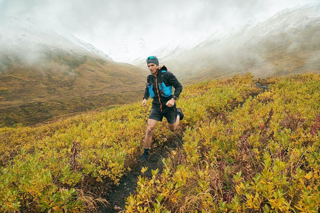 A male trail runner in a blue vest runs uphill on a path lined with vegetation. The background features mountains under a cloudy sky.