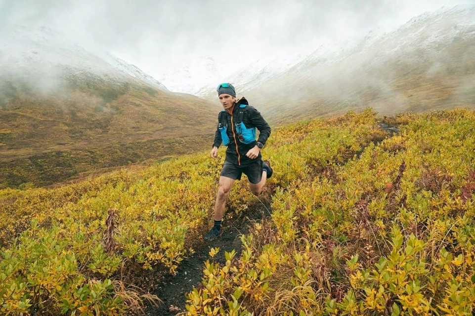 A male trail runner in a blue vest runs uphill on a path lined with vegetation. The background features mountains under a cloudy sky.