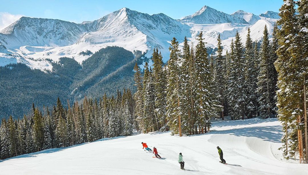 Scenic mountain landscape with snow-covered peaks, tall evergreen trees, and skiers descending a slope on a sunny day.