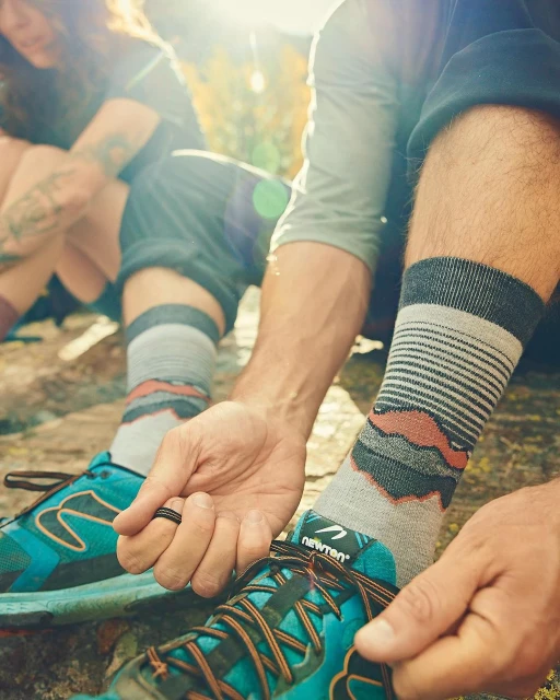 Close-up of a person tying bright blue and orange hiking shoes with patterned grey socks outdoors with a second person in the background.