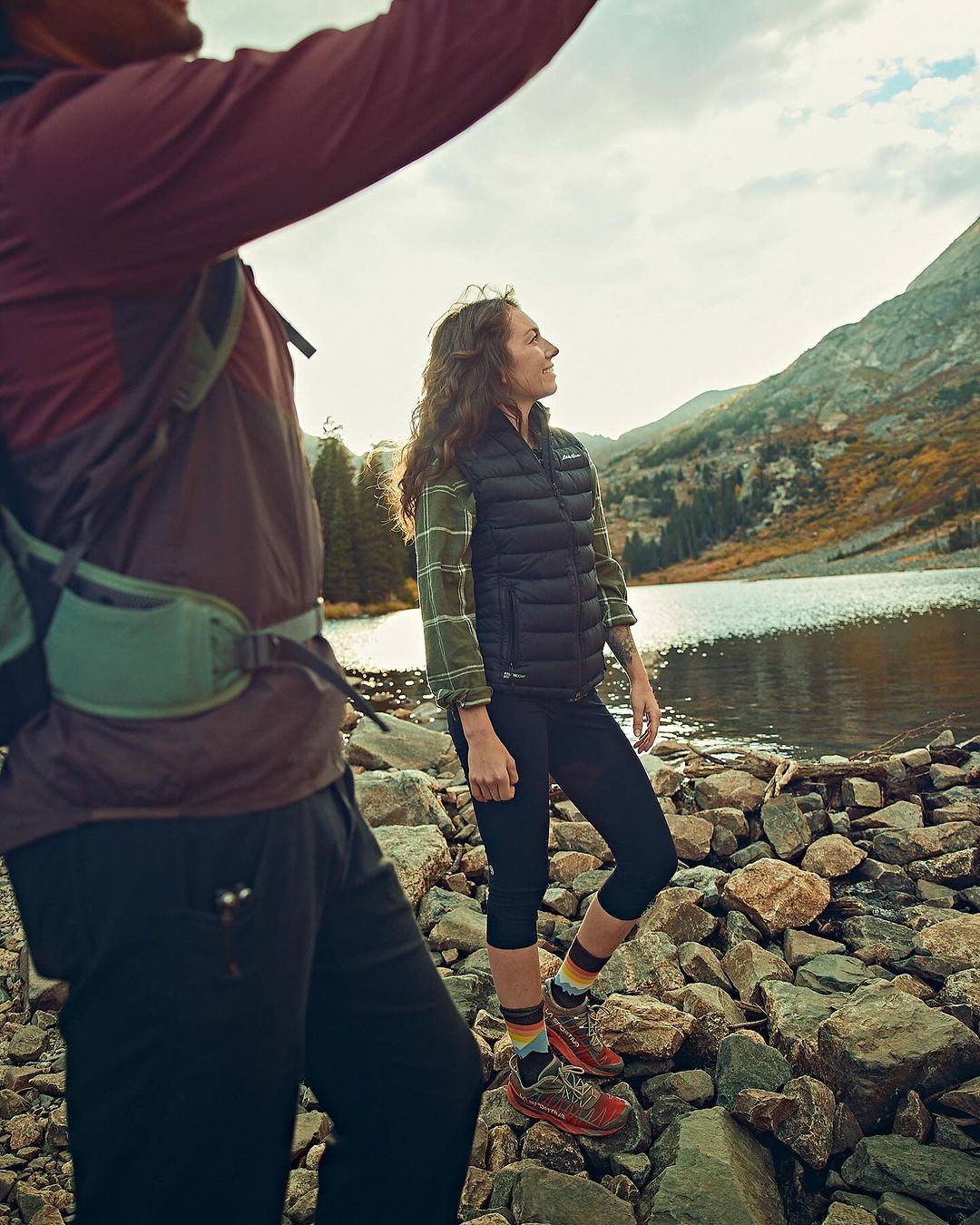 Female hiker standing on rocks, mountain and lake backdrop on a sunny day. A male hiker is partially in the foreground.