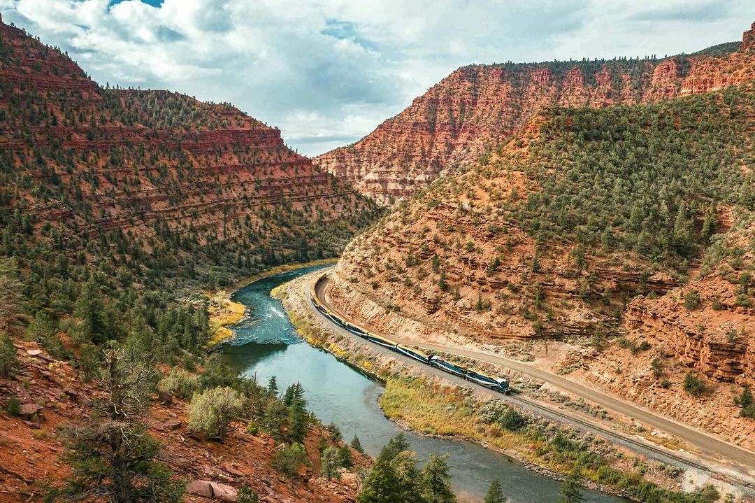 Scenic landscape featuring a train traveling along tracks beside a river winding through a canyon with red rock cliffs and green trees.
