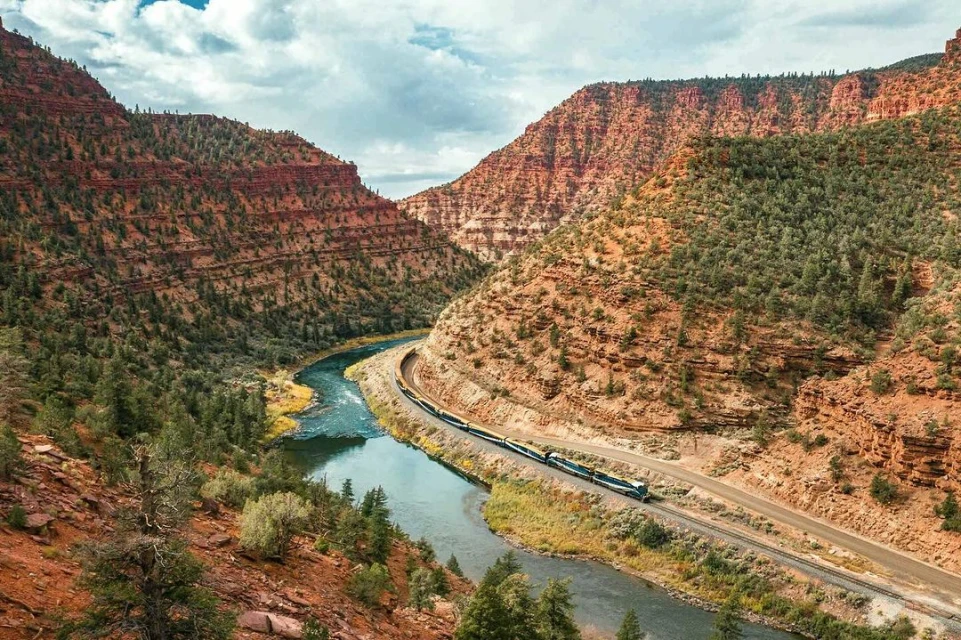 Scenic landscape featuring a train traveling along tracks beside a river winding through a canyon with red rock cliffs and green trees.