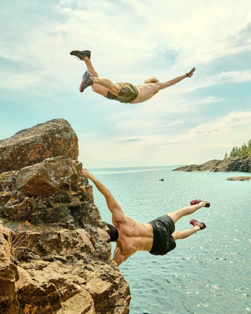 Two shirtless men leap from a rocky cliffside into a blue lake under a partly cloudy sky, showcasing athleticism and summer adventure.