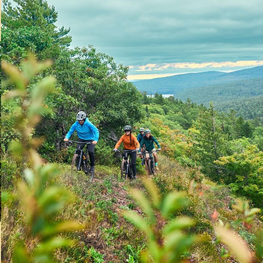 A group of four people riding mountain bikes on a dirt trail through a forest, with mountains and a lake visible in the distance under a cloudy sky.